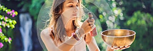 Woman playing on Tibetan singing bowl while sitting on yoga mat against a waterfall. Vintage tonned. Beautiful girl with