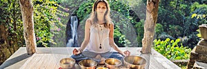 Woman playing on Tibetan singing bowl while sitting on yoga mat against a waterfall. Vintage tonned. Beautiful girl with