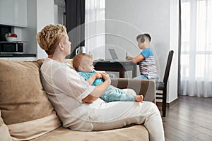 Woman playing with toddler while eldest son using laptop