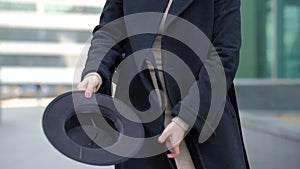 Woman plays with modern black hat moving wide-brimmed chapeau at black coat bottom in street slow motion low angle shot.