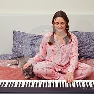 A woman plays the piano sitting in a home bedroom next to a cat