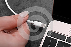 Woman plugging USB cable with lightning connector into laptop port on dark table, closeup
