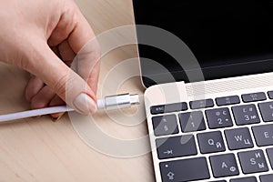 Woman plugging USB cable with type C connector into laptop port on light wooden table, closeup