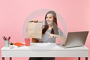 Woman pointing on brown clear empty blank craft paper bag, work at office with laptop isolated on pink background. Food