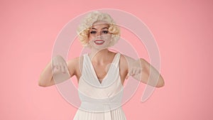 A woman points down with her index fingers. Woman in the image of Marilyn Monroe in the studio on a pink background