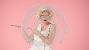 The woman points her hand and index finger to the side. Woman in the image of Marilyn Monroe in the studio on a pink