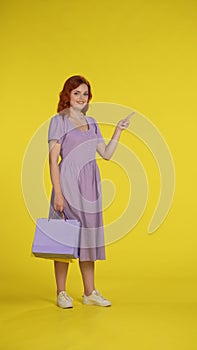 A woman points to something with her index finger to the side. Full length woman with shopping bags in studio on yellow