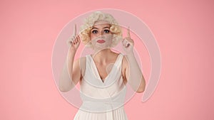 Woman points upward with her index fingers. Woman in white dress and wig, in the look of Marilyn Monroe in studio on