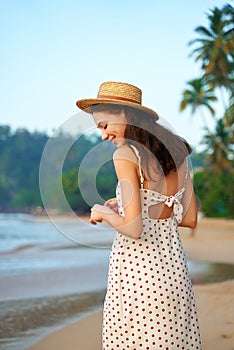 Woman in polka dot dress enjoys sunrise on sandy beach. Boho straw hat, vintage summer style. Solo traveler cherishes