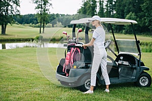woman in polo and cap with golf gear standing at golf cart at golf course