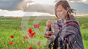 Woman in poncho with singing bowl sits on flower field under cloudy sunset sky.