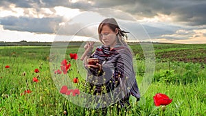 Woman in poncho with singing bowl sits on flower field under cloudy sunset sky.