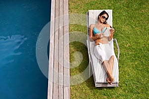 Woman, pool and relax on chair with book or novel for tanning on holiday or resort in summer and sunshine. Young person