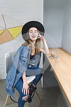 Woman poses to the camera in a cozy cafe for a cup of hot drink near the window. Portrait of a hipsters girl in a cafe.