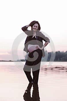 Woman poses in water. Pregnant young Asian woman in dark suit stands in water on seashore at sunset and holds her long