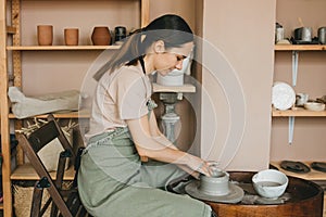 Woman pottery maker works in clay studio on pottery wheel against shelves with vases and pots.