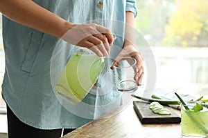 Woman pouring aloe vera cocktail from jug into glass at wooden table