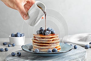 Woman pouring chocolate syrup onto fresh pancakes with blueberries at grey table