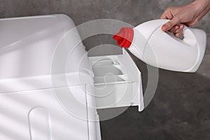 Woman pouring fabric softener from bottle into washing machine near grey wall, closeup