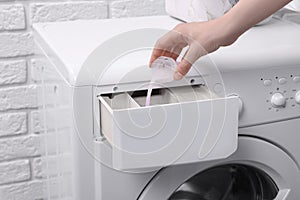 Woman pouring fabric softener from cap into washing machine near white brick wall, closeup