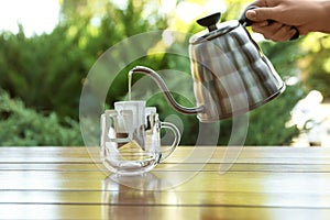 Woman pouring hot water into glass cup with drip coffee bag from kettle at wooden table, closeup