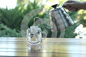 Woman pouring hot water into glass cup with drip coffee bag from kettle at wooden table, closeup