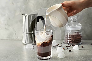 Woman pouring milk into glass with cold coffee on grey table