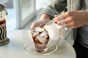 Woman pouring milk into glass cup with cold coffee at light table