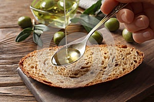 Woman pouring olive oil into spoon over bread at wooden table, closeup