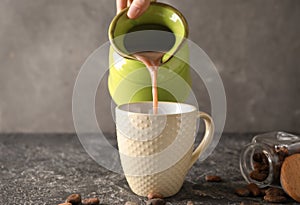 Woman pouring tasty cocoa from jug into cup on grunge table