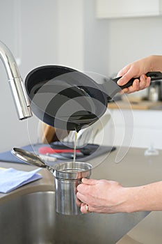 Woman pouring used cooking oil from an old frying pan into a metal cooking container in her kitchen
