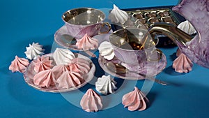 A woman pours tea into beautiful pink porcelain cups that stand on a blue background. Next to them is a saucer of sweets