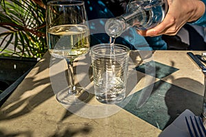 A woman pours water into a glass next to a glass of white wine i photo