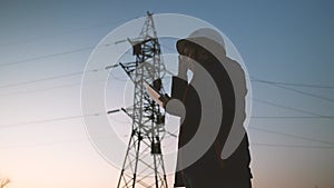 A woman power engineer in white helmet inspects power line using data from electrical sensors on a tablet. High voltage