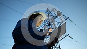A woman power engineer in white helmet inspects power line using data from electrical sensors on a tablet. High voltage