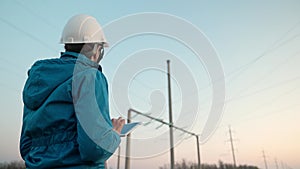 A woman power engineer in white helmet inspects power line using data from electrical sensors on a tablet. High voltage