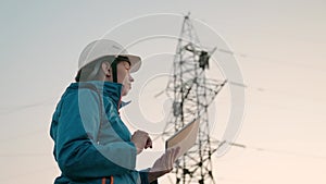 A woman power engineer in white helmet inspects power line using data from electrical sensors on a tablet. High voltage