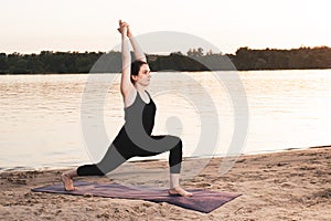 A woman practices yoga while standing in the position of Warrior 1 or Virabhadrasana One. Yoga at sunset in nature near