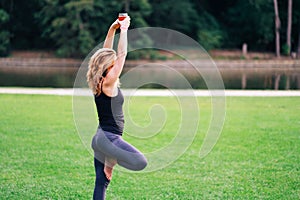A woman practicing drunk yoga outdoor at the park. Woman holding a glass of alcohol doing pilates