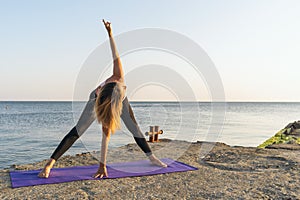 A woman practicing yoga doing the Parivritta Padottanasana twisting exercise while standing on the pier on a sunny summer evening