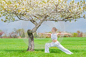 Woman is practicing yoga, doing Virabhadrasana exercise, standing in Warrior pose near tree