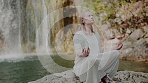 Woman Practicing Yoga Exercises Near Fresh Waterfall In Mountains