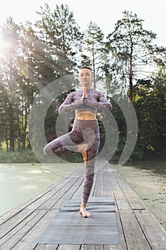 A woman practicing yoga, in the morning on a wooden bridge in the park, performs the vrikshasana exercise, tree pose