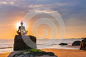 Woman is practicing yoga sitting on stone in Lotus pose at sunset. Silhouette of woman meditating on the beach