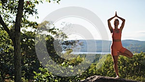 Woman is practicing yoga in tree pose, at a beautiful exotic place on the top of a mountain at sunset. The camera moves from left