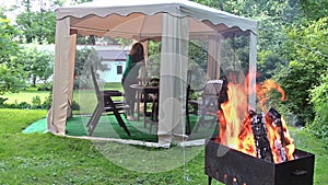 Woman prepare supper table in outdoor bower. Firewood burn