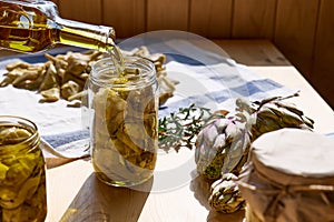 Woman prepares canned artichokes and pours oil into the glass jar with artichokes and herbs. Homemade healthy eating.