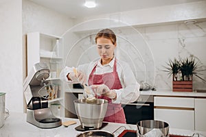 Woman prepares dough using kitchen mixer, pouring ingredients into steel bowl. Cooking homemade cake