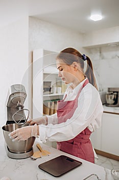 Woman prepares dough using kitchen mixer, pouring ingredients into steel bowl. Cooking homemade cake