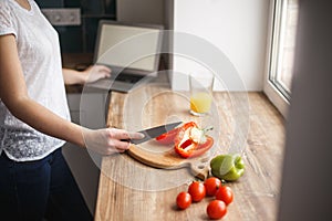 A woman is preparing a meal and working on a laptop. Nearby is a glass of juice. Sliced peppers and whole tomatoes